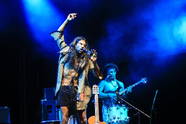 Un chanteur aux cheveux longs lève le bras en chantant au micro sur une scène de concert illuminée de bleu.