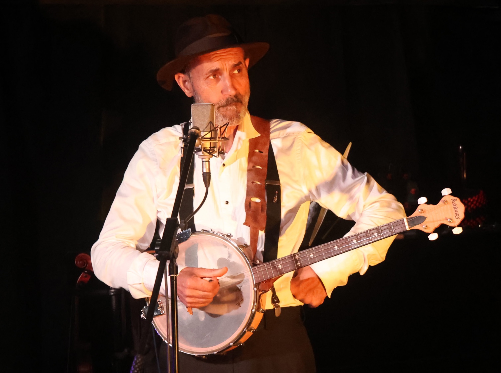 Musicien barbu coiffé d'un chapeau et portant des bretelles jouant du banjo devant un micro sur scène.