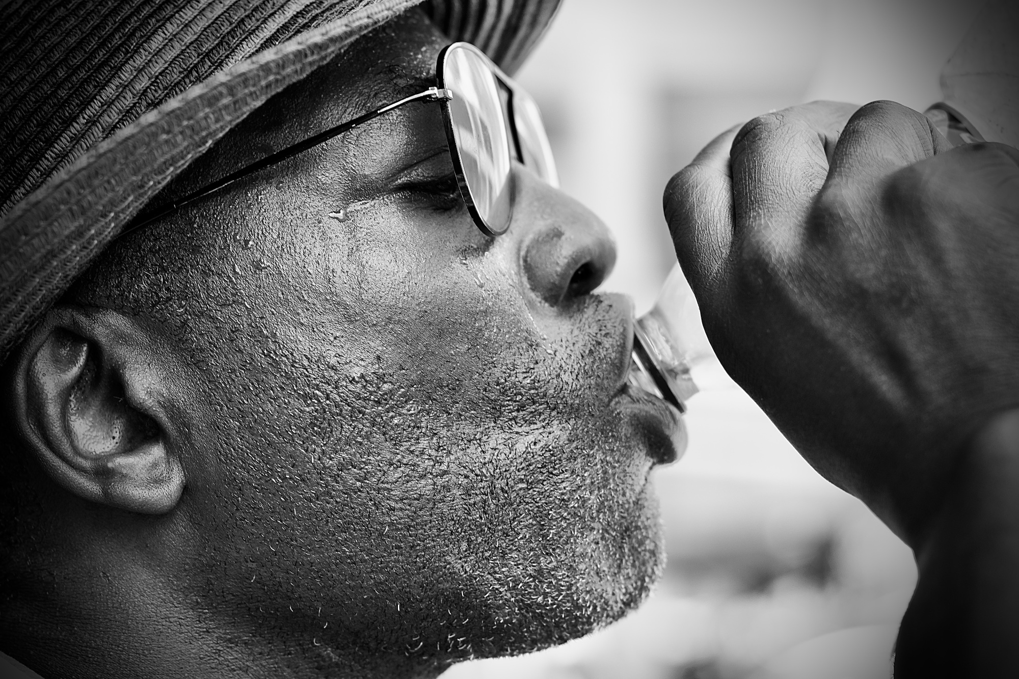 Gros plan en noir et blanc de profil d'un homme portant un chapeau et des lunettes de soleil qui boit à une bouteille.