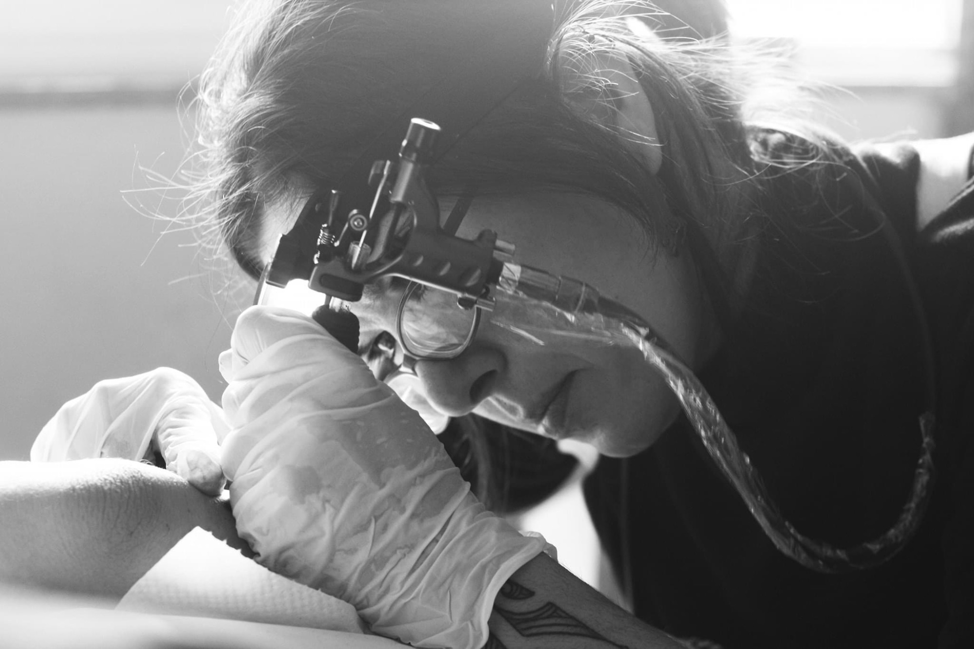 Gros plan en noir et blanc d'une tatoueuse gantée et concentrée utilisant sa machine à tatouer sur le bras d'un client.