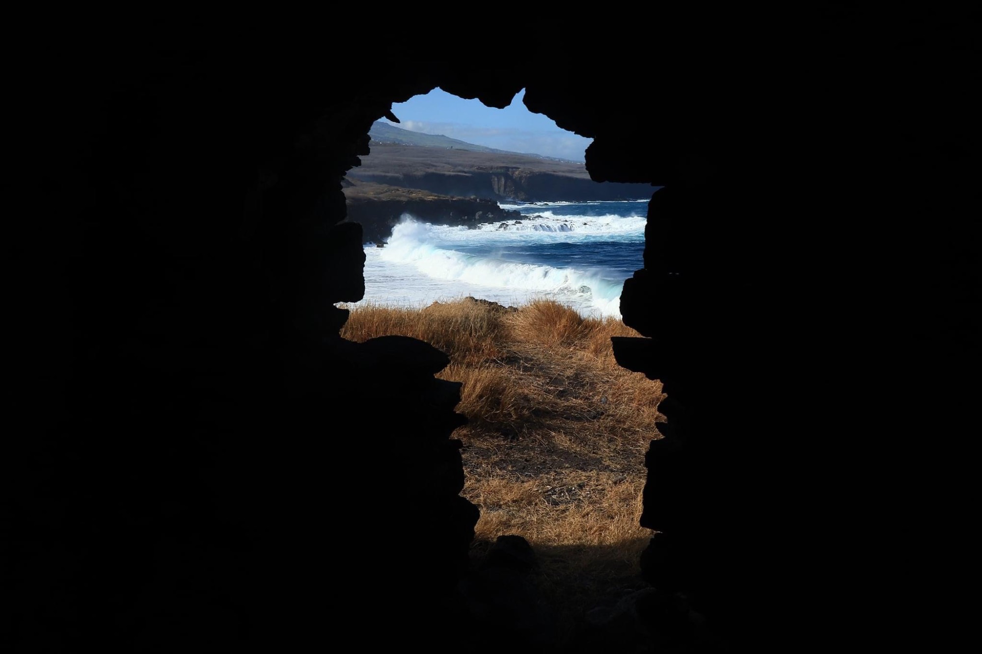 Vue d'une côte sauvage et de vagues déferlantes à travers une ouverture sombre dans un mur de pierre.