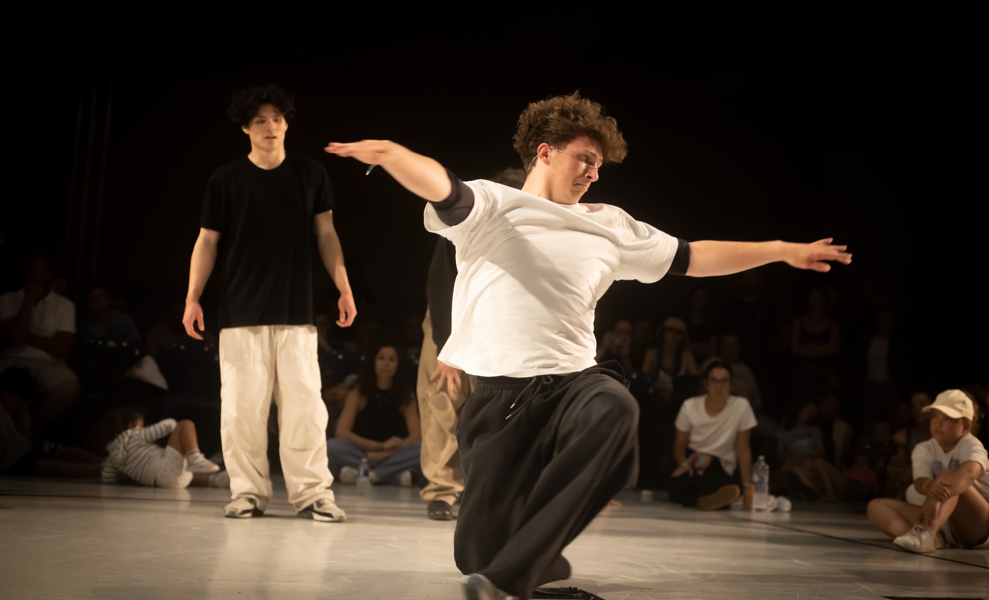 Un jeune danseur en t-shirt blanc et pantalon large en plein mouvement sur scène devant un public assis.